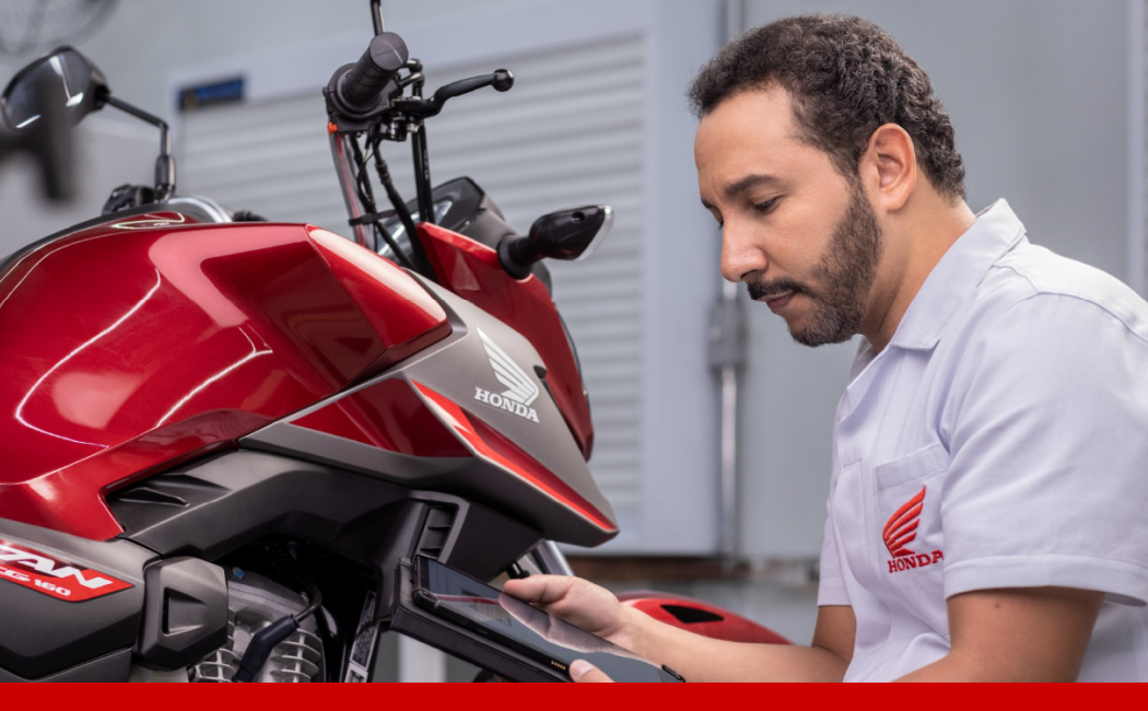 Técnico da Honda utilizando um tablet para realizar diagnóstico ou atendimento em uma motocicleta vermelha dentro de uma oficina.