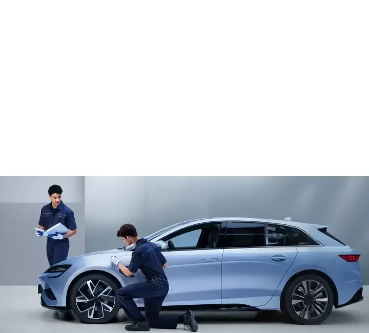 Dois técnicos automotivos vestindo uniformes azuis-escuros e luvas brancas realizam a inspeção de um carro moderno de cor azul-claro em um ambiente de oficina limpo e minimalista. Um dos técnicos está ajoelhado ao lado da roda dianteira, examinando detalhadamente o para-lama do veículo. O outro técnico está de pé logo atrás, segurando uma prancheta branca e acompanhando o procedimento. O carro é exibido de perfil, destacando suas rodas de liga leve com design aerodinâmico.
