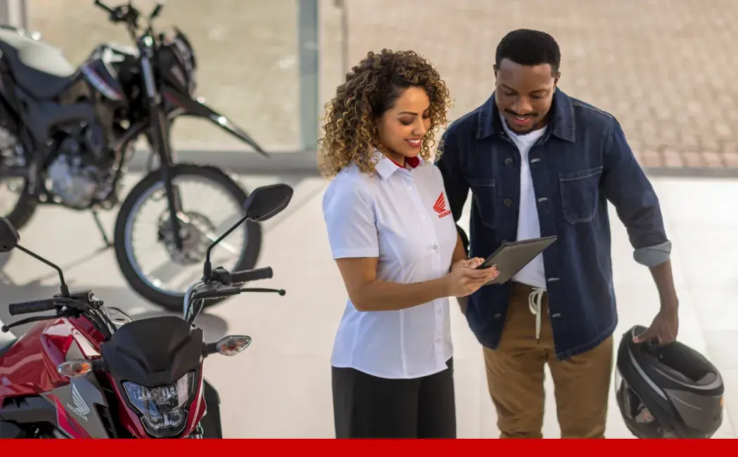 Da esquerda para a direita, um homem, uma mulher e outro homem vestindo uniformes da Honda Motos dentro de uma concessionária com motos ao fundo e ao redor