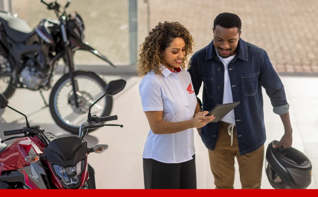 Da esquerda para a direita, um homem, uma mulher e outro homem vestindo uniformes da Honda Motos dentro de uma concessionária com motos ao fundo e ao redor