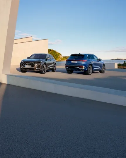 Audi Q5 cinza e Audi SQ5 azul estacionados diante de uma construção de concreto à beira-mar