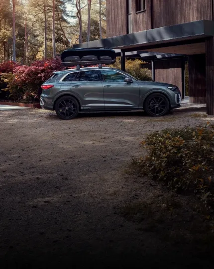 SUV Audi cinza com bagageiro de teto, estacionado em frente a uma casa cercada por floresta de outono