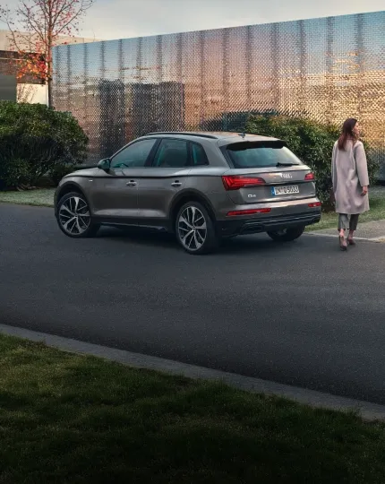 Audi Q5 marrom estacionado em rua arborizada, com uma mulher caminhando ao lado