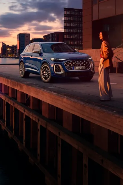 Audi SQ5 azul estacionado em um cais ao pôr do sol, com o skyline da cidade ao fundo e uma mulher ao lado