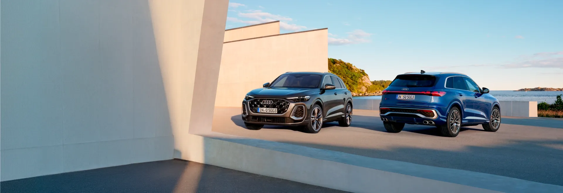 Audi Q5 cinza e Audi SQ5 azul estacionados diante de uma construção de concreto à beira-mar
