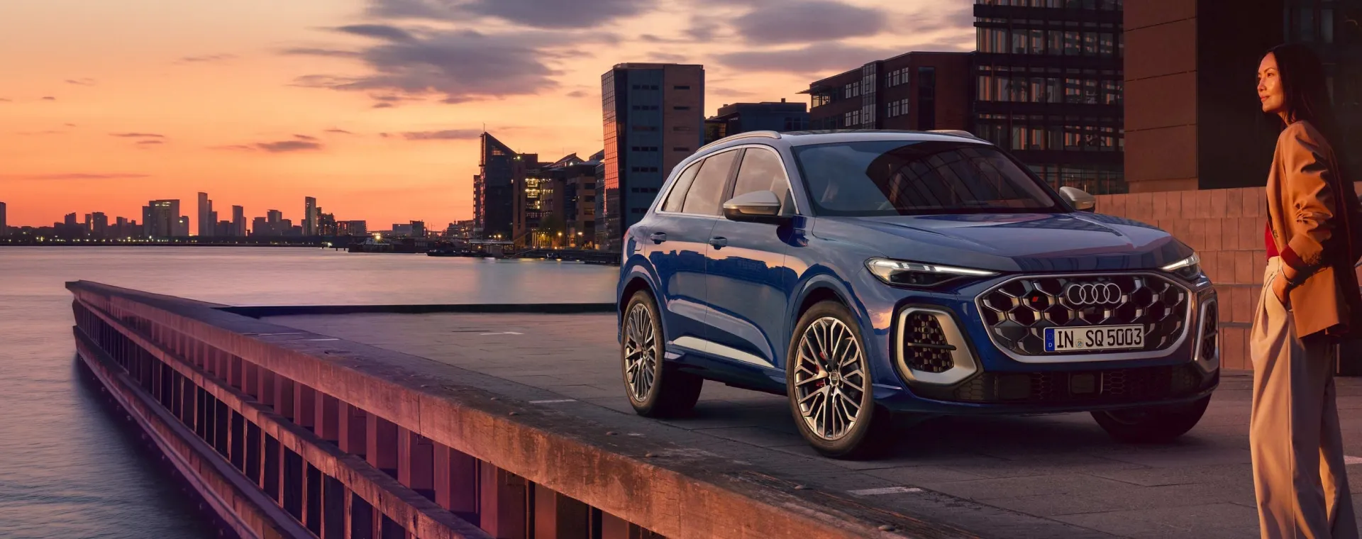 Audi SQ5 azul estacionado em um cais ao pôr do sol, com o skyline da cidade ao fundo e uma mulher ao lado