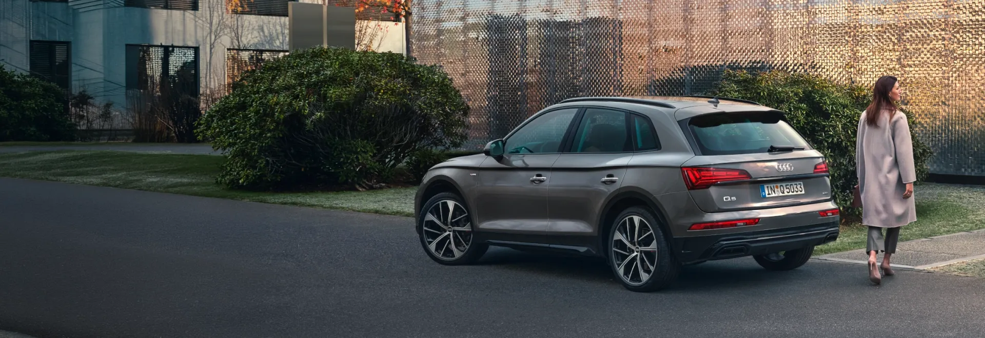 Audi Q5 marrom estacionado em rua arborizada, com uma mulher caminhando ao lado