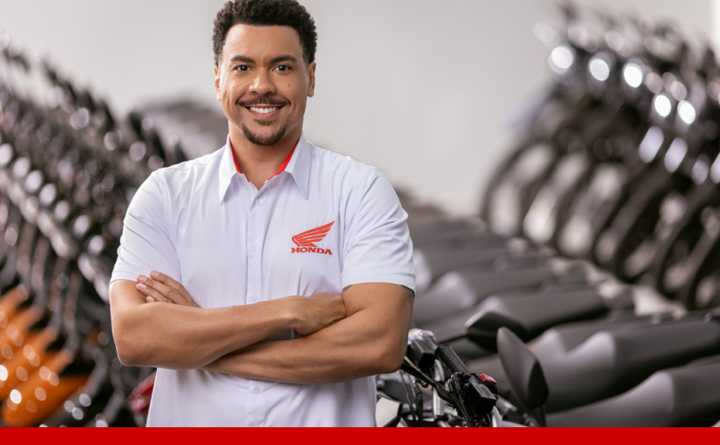Funcionário da Honda sorrindo com os braços cruzados em uma concessionária, com motocicletas ao fundo.